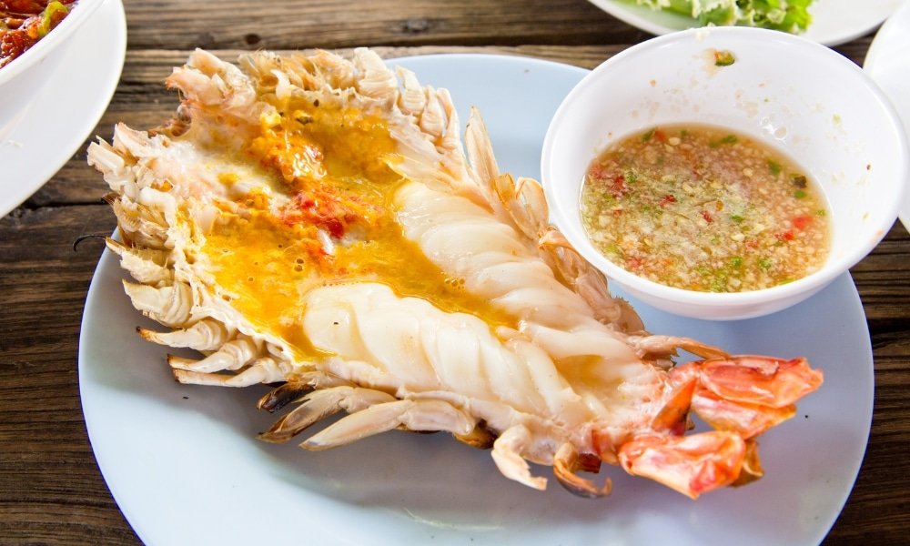 กุ้งแม่น้ำย่างผ่าครึ่งวางบนจานข้างชามน้ำจิ้มรสเผ็ด—เมนูห้ามพลาดเมื่อคุณมาเที่ยวอยุธยาและสำรวจรสชาติท้องถิ่นที่สถานที่ท่องเที่ยวแห่งนี้ในอยุธยา.
