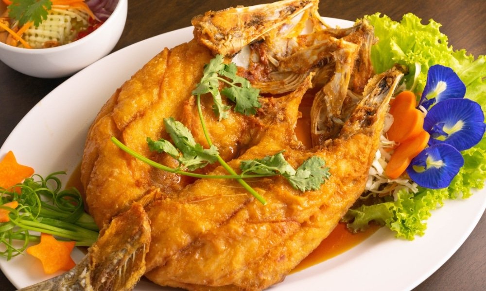 ปลาทอดเสิร์ฟบนจานขาวพร้อมผักกาดหอม, แครอทหั่น, ดอกไม้ที่กินได้, และผักชี—เหมาะสำหรับการเพลิดเพลินหลังจากสำรวจ ที่เที่ยวอยุธยา, พร้อมชามผักอยู่ด้านหลัง.