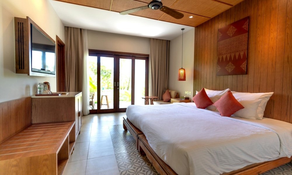 ห้องพักโรงแรมหัวหินสไตล์โมเดิร์น พร้อมเตียงขนาดใหญ่ เฟอร์นิเจอร์ไม้ ทีวีติดผนัง และประตูกระจกเปิดออกสู่ลานระเบียงที่รายล้อมด้วยต้นไม้สีเขียว เหมาะสำหรับผู้ที่มองหาที่พักหัวหินที่มีสไตล์และความสะดวกสบาย.