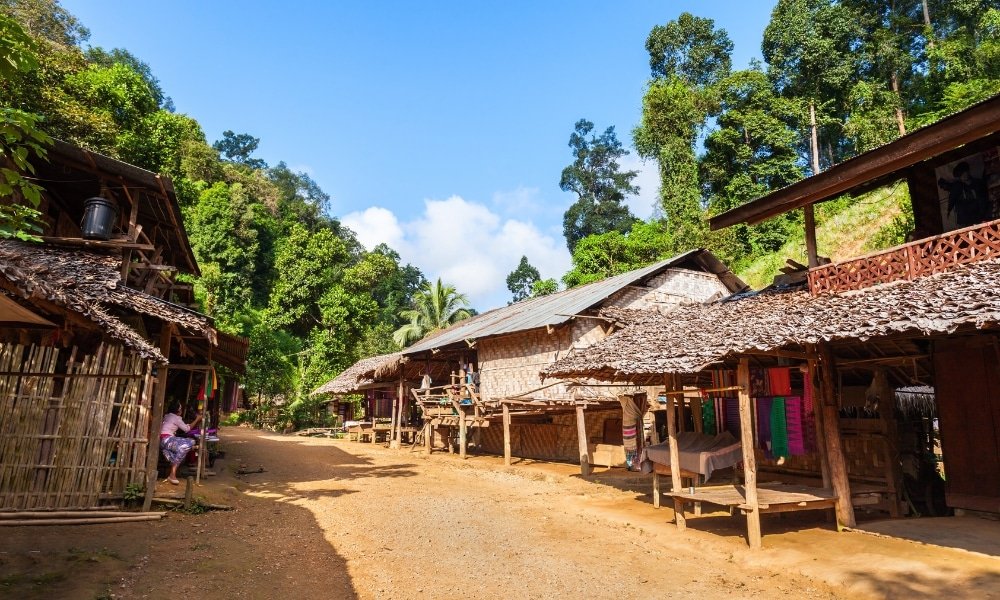 ภาพหมู่บ้านชนบทใน ที่เที่ยวอยุธยา พร้อมบ้านไม้แบบดั้งเดิม หลังคามุงจาก และถนนลูกรังที่ล้อมรอบด้วยต้นไม้เขียวชอุ่มภายใต้ท้องฟ้าสีคราม.