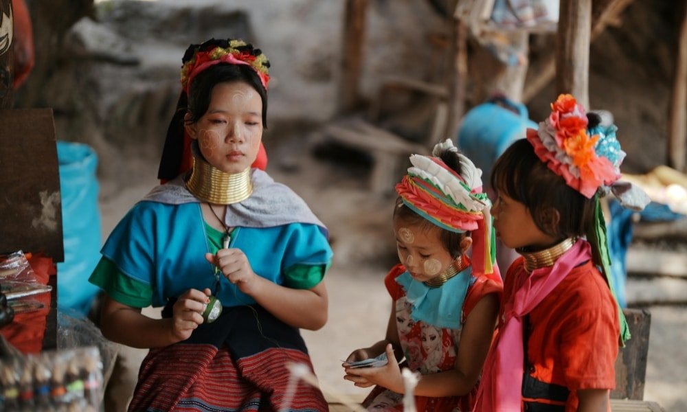 ผู้หญิงคนหนึ่งและเด็กหญิงสองคนจากเผ่ากะยาอานั่งอยู่กลางแจ้งในชุดประจำเผ่าและสร้อยคอทองเหลืองที่คล้องรอบคอของพวกเธอ โดยมีสิ่งของต่าง ๆ อยู่รอบตัว—เป็นภาพวัฒนธรรมที่น่าสนใจสำหรับผู้ที่มาเยือนสถานที่ท่องเที่ยวในอยุธยา.