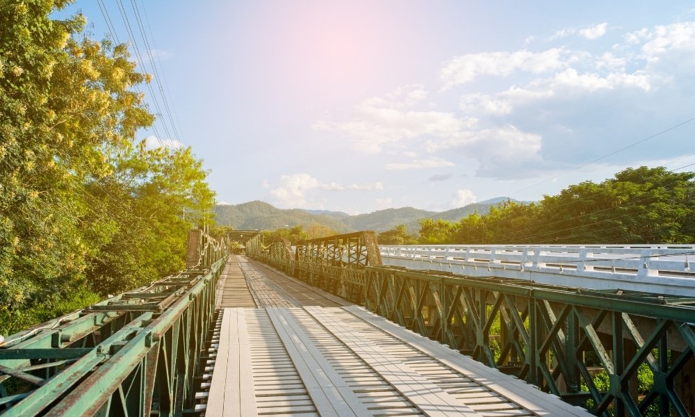 สะพานโครงเหล็กที่มีพื้นไม้ทอดข้ามพื้นที่สีเขียวในอยุธยาที่เที่ยว โดยมีสะพานอีกแห่งที่คล้ายกันอยู่ข้างๆ ภายใต้ท้องฟ้าที่แจ่มใส มีเมฆและภูเขาอยู่ไกลๆ.