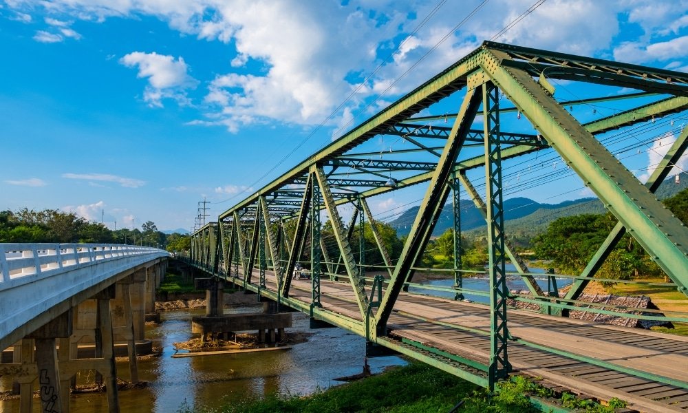 สะพานโครงเหล็กสีเขียวทอดข้ามแม่น้ำข้างสะพานคอนกรีตสมัยใหม่ ท่ามกลางภูเขาและต้นไม้ภายใต้ท้องฟ้าสีคราม—ทัศนียภาพที่สะดุดตา เหมาะสำหรับผู้ที่ต้องการสำรวจสถานที่ท่องเที่ยวในอยุธยาหรือวางแผนการผจญภัยครั้งต่อไปในอยุธยา.