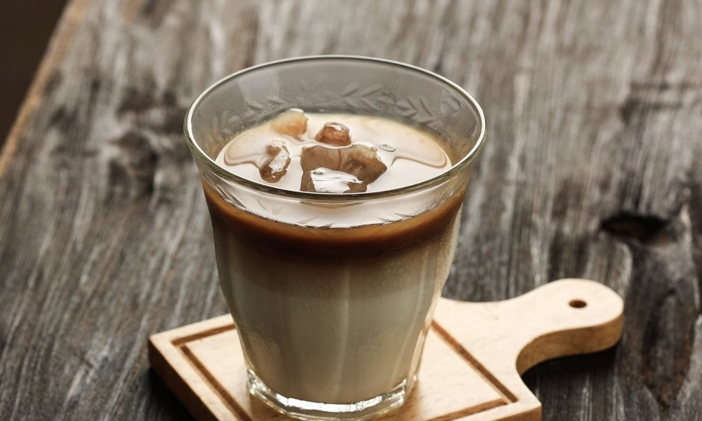 กาแฟเย็นใส่นมแก้วหนึ่งวางอยู่บนที่รองแก้วไม้บนโต๊ะไม้สไตล์ชนบท—เหมาะสำหรับการผ่อนคลายหลังจากวันแห่งการสำรวจสถานที่ท่องเที่ยวเชียงใหม่.