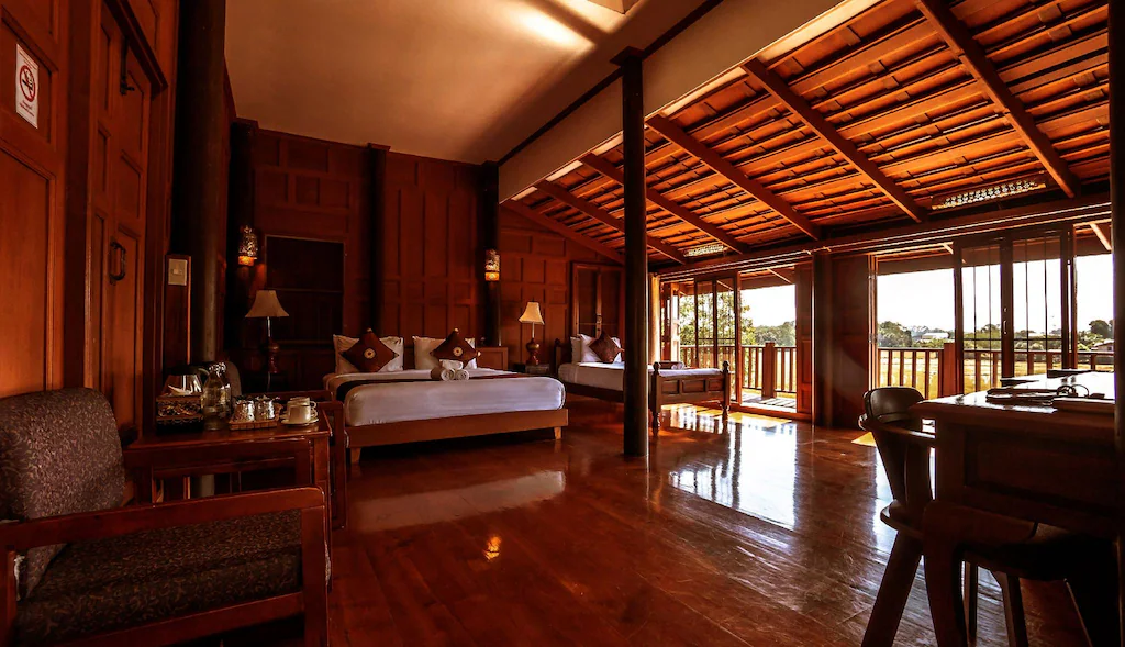 ห้องพักโรงแรมกว้างขวางพร้อมพื้นไม้ เตียงสองเตียง พื้นที่นั่งเล่น และหน้าต่างบานใหญ่ที่รับแสงธรรมชาติพร้อมวิวระเบียง—เหมาะสำหรับผู้ที่มองหาที่พักในน่านสำหรับการเดินทาง.