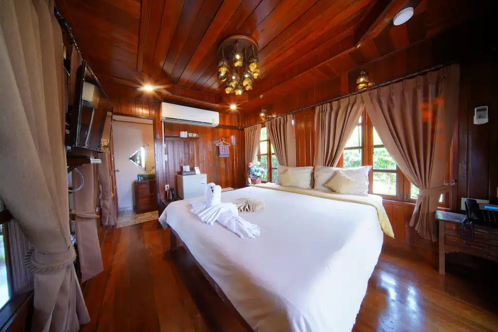 ห้องนอนไม้แสนอบอุ่นที่ ที่พักอยุธยา พร้อมเตียงขนาดใหญ่, ศิลปะผ้าขนหนู, ทีวี, เครื่องปรับอากาศ, ม่านหน้าต่าง, และแสงสว่างอบอุ่นเพื่อการพักผ่อนที่ผ่อนคลาย.