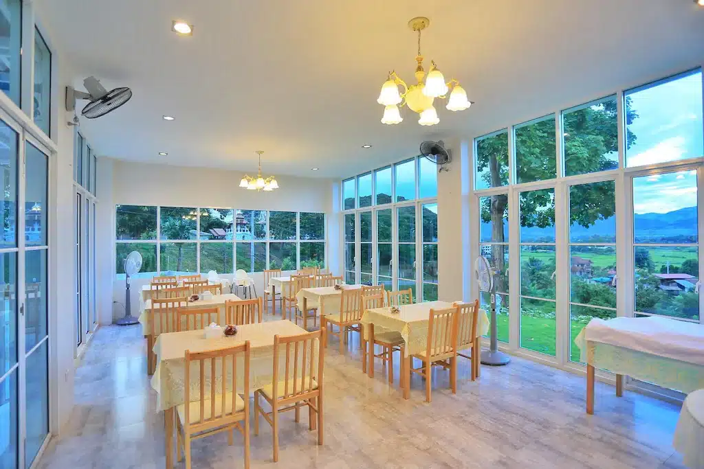 พื้นที่รับประทานอาหารสว่างสดใสพร้อมโต๊ะและเก้าอี้ไม้ หน้าต่างกระจกบานใหญ่ที่มองเห็นวิวต้นไม้และภูเขาด้านนอก เหมาะสำหรับการพักผ่อนที่ที่พักปัว พร้อมไฟเพดานและพัดลมที่ช่วยเสริมบรรยากาศอบอุ่น.