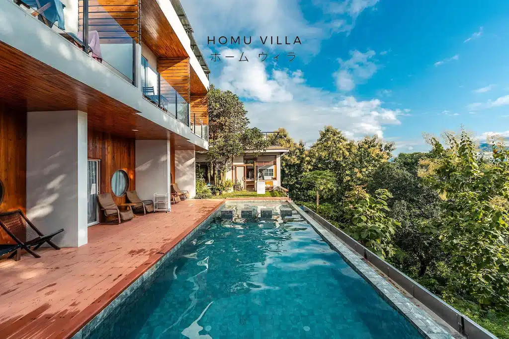 วิลล่าสมัยใหม่พร้อมตกแต่งด้วยไม้ สระว่ายน้ำอินฟินิตี้กลางแจ้ง เก้าอี้อาบแดด และพื้นที่สีเขียวชอุ่มภายใต้ท้องฟ้าสีคราม—ที่พักในน่านที่สมบูรณ์แบบสำหรับการพักผ่อนครั้งต่อไปของคุณ ข้อความระบุว่า "HOMU VILLA" เป็นภาษาอังกฤษและภาษาญี่ปุ่น.