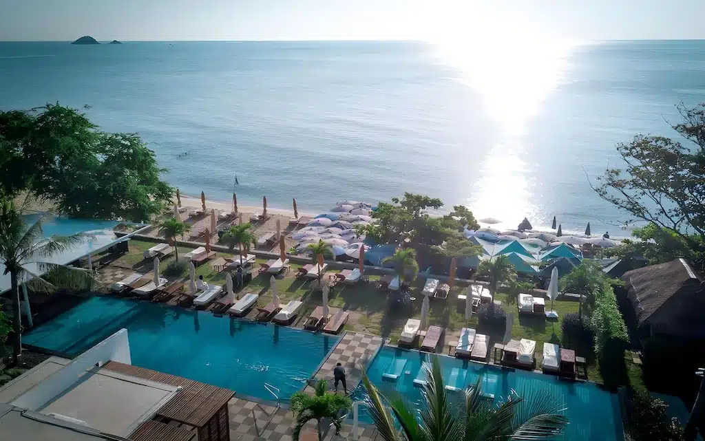 ภาพมุมสูงของรีสอร์ทริมชายหาดและสระว่ายน้ำที่ล้อมรอบด้วยต้นปาล์มและเก้าอี้อาบแดดในยามพระอาทิตย์ตก—เหมาะสำหรับผู้ที่มองหาที่พักหัวหินติดทะเล หรือโรงแรมหัวหินที่มีวิวทะเลสวยงามและบรรยากาศผ่อนคลาย.