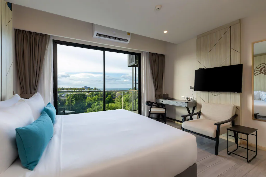 ห้องพักโรงแรมสมัยใหม่ที่ โรงแรมหัวหิน พร้อมเตียงขนาดใหญ่ โต๊ะทำงาน เก้าอี้นวม ทีวีติดผนัง และระเบียงที่มองเห็นวิวสีเขียวและภูเขาไกลภายใต้ท้องฟ้าที่มีเมฆบางส่วน เป็นตัวเลือกที่สมบูรณ์แบบสำหรับ ที่พักหัวหินราคาถูก.