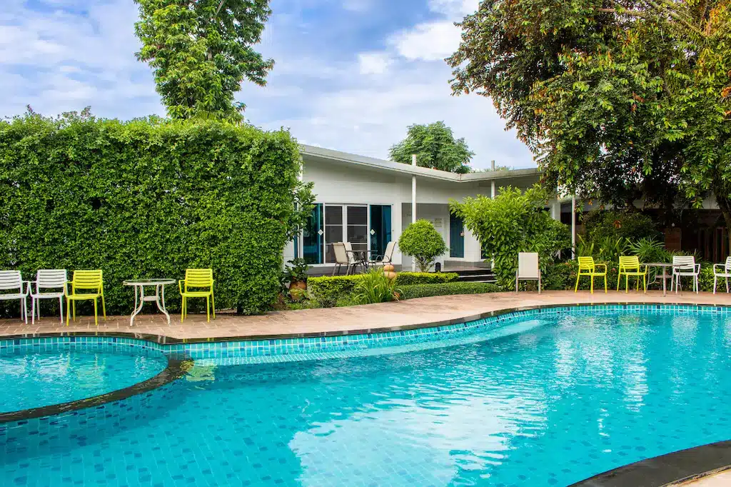 สระว่ายน้ำกลางแจ้งที่ใสสะอาดพร้อมขอบกระเบื้อง ล้อมรอบด้วยโต๊ะและเก้าอี้ลานกลางแจ้ง พืชสีเขียว และอาคารสีขาวชั้นเดียวในพื้นหลัง—เหมาะสำหรับการพักผ่อนที่ ที่พักบ่อเกลือ หรือที่พักในน่านครั้งต่อไปของคุณ.