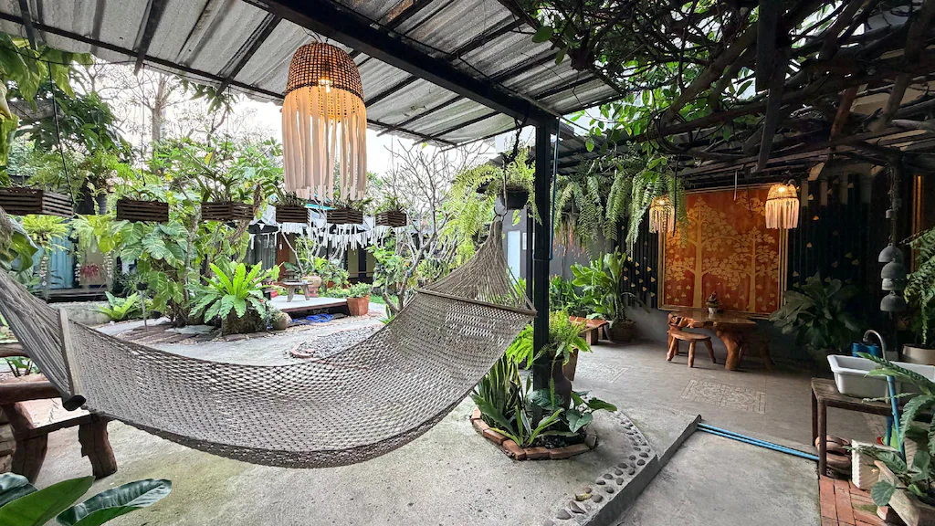 ระเบียงที่มีหลังคาคลุมที่ ที่พักอยุธยา แห่งนี้มีเปลญวนที่ถักทอ โคมไฟแขวน ต้นไม้ในกระถาง เฟอร์นิเจอร์ไม้ และพืชพรรณเขียวชอุ่มเพื่อสร้างบรรยากาศสวนที่เงียบสงบ.