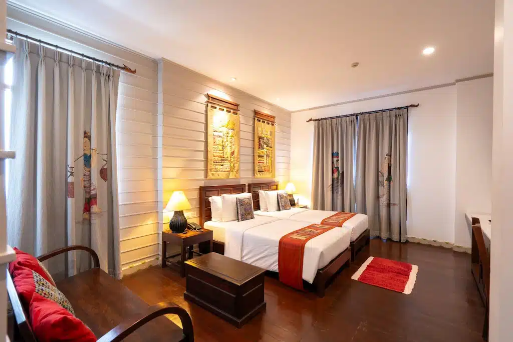 ห้องพักโรงแรมพร้อมเตียงเดี่ยวสองเตียง พื้นไม้ พื้นที่นั่งเล่น หมอนตกแต่ง โคมไฟ ม่าน และงานศิลปะบนผนัง ส่องสว่างอย่างอบอุ่น—เหมาะอย่างยิ่งสำหรับนักเดินทางที่กำลังมองหาที่พักในน่านหลังจากสำรวจเมือง.