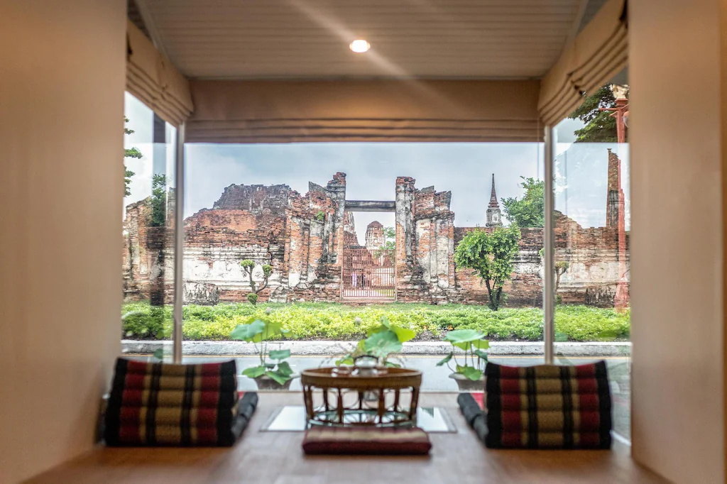 วิวจากหน้าต่างของห้องพักที่แสนสบายในอยุธยา พร้อมด้วยหมอนและโต๊ะเล็ก ๆ มองเห็นซากปรักหักพังของอิฐโบราณและต้นไม้เขียวชอุ่มด้านนอก.