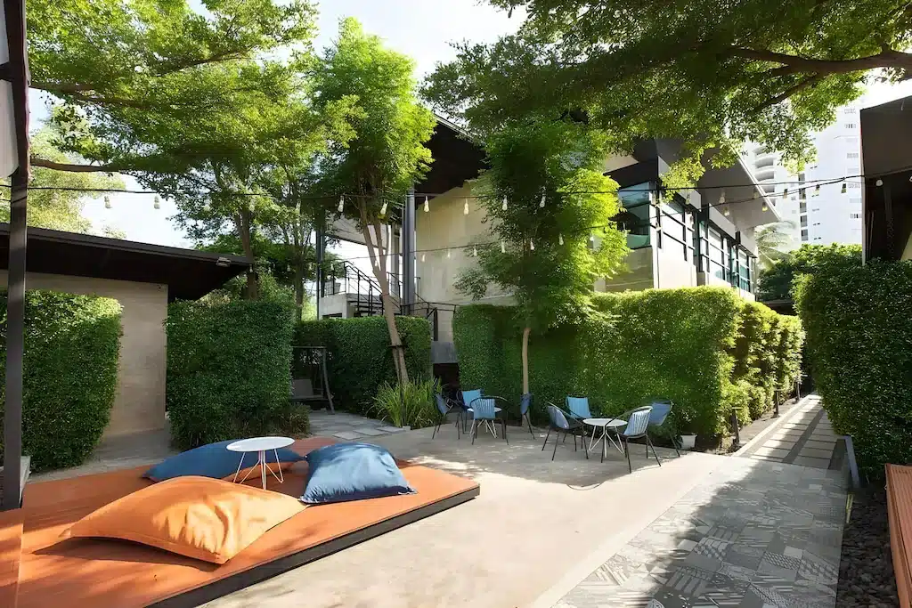 ลานระเบียงกลางแจ้งสมัยใหม่พร้อมเบาะรองนั่งขนาดใหญ่ โต๊ะเตี้ย เก้าอี้สีฟ้า และพืชพรรณเขียวชอุ่ม รวมถึงรั้วต้นไม้และต้นไม้ใหญ่—เหมาะสำหรับการพักผ่อนที่ ที่พักหัวหิน หรือ โรงแรมหัวหิน ท่ามกลางตึกสูงในเมืองเป็นฉากหลัง.