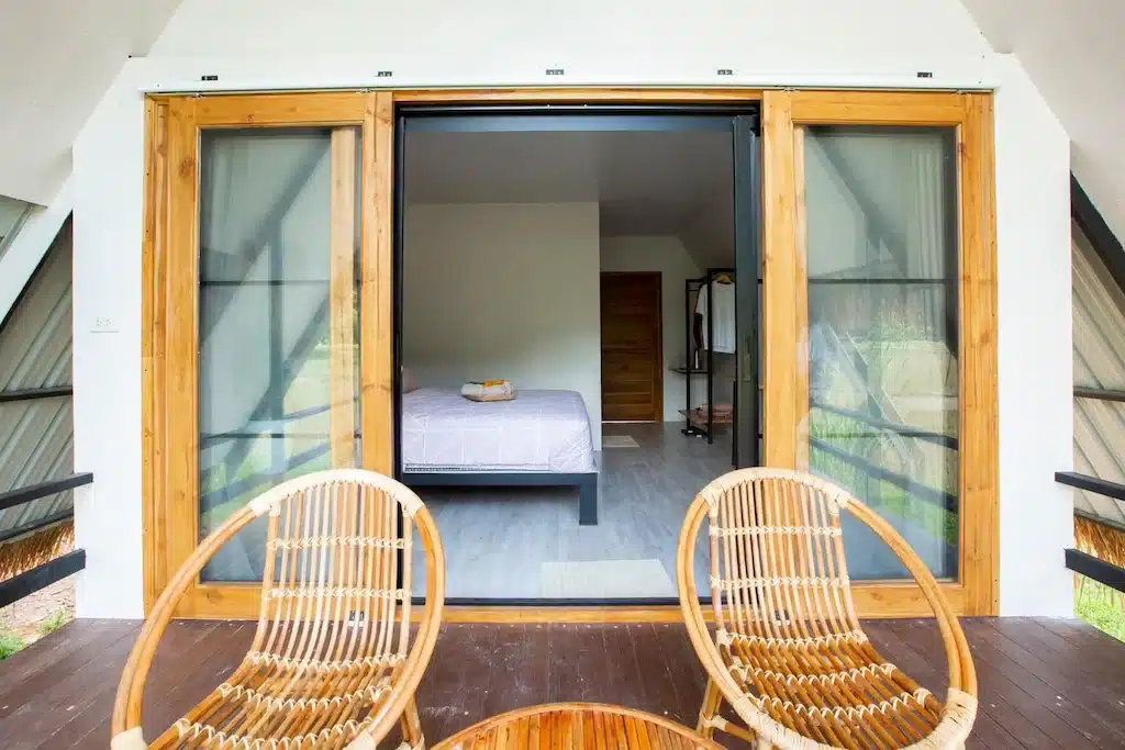 วิวจากระเบียงไม้พร้อมเก้าอี้หวายสองตัว มองเข้าไปในห้องนอนที่มีเตียงคู่ ผนังสีขาว ประตูไม้ และผ้าขนหนูวางอยู่บนเตียง—เหมาะสำหรับการพักผ่อนที่ ที่พักบ่อเกลือ ใน น่าน.