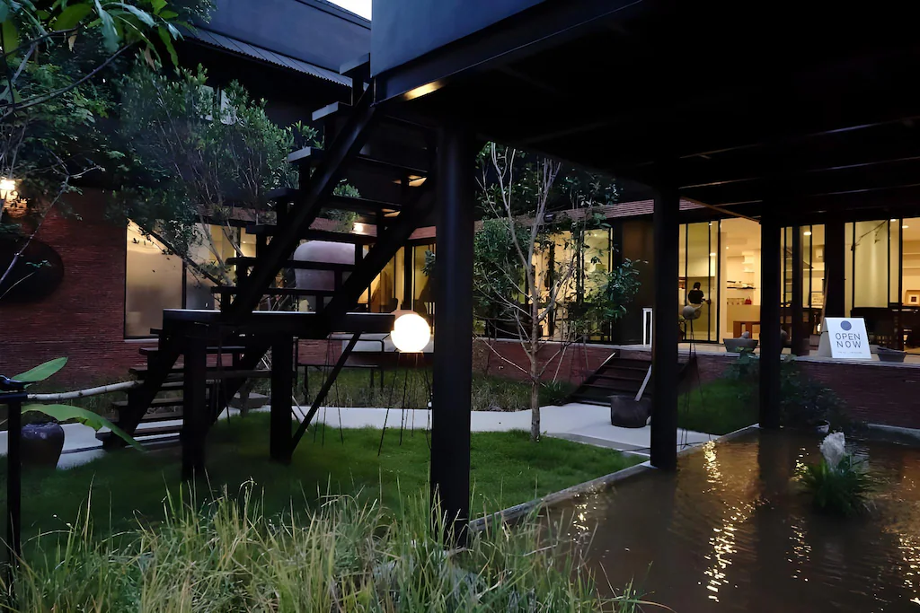 อาคารสมัยใหม่พร้อมหน้าต่างบานใหญ่ บันไดภายนอก ลานหญ้า และสระน้ำยามพลบค่ำ; ป้ายที่ติดอยู่หน้าต่างเขียนว่า "เปิดแล้ว" ตัวเลือกที่สมบูรณ์แบบสำหรับ ที่พักอยุธยา ใกล้แม่น้ำ—การพักผ่อนที่สมบูรณ์แบบในบรรยากาศที่มีสไตล์.