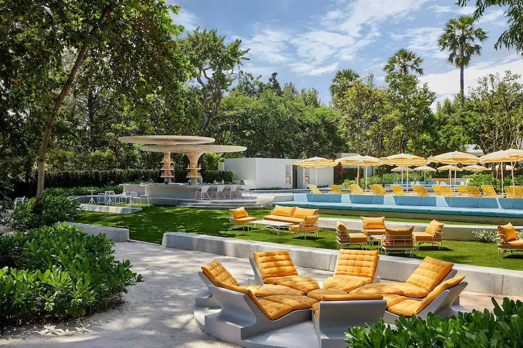 บริเวณสระว่ายน้ำกลางแจ้งที่โรงแรมหัวหิน พร้อมเก้าอี้เลานจ์สีเหลือง ร่ม น้ำพุ สนามหญ้าสีเขียว และต้นไม้ล้อมรอบภายใต้ท้องฟ้าที่มีเมฆบางส่วน—ที่พักหัวหินที่เหมาะสำหรับผู้ที่มองหาที่พักหัวหินราคาถูก.