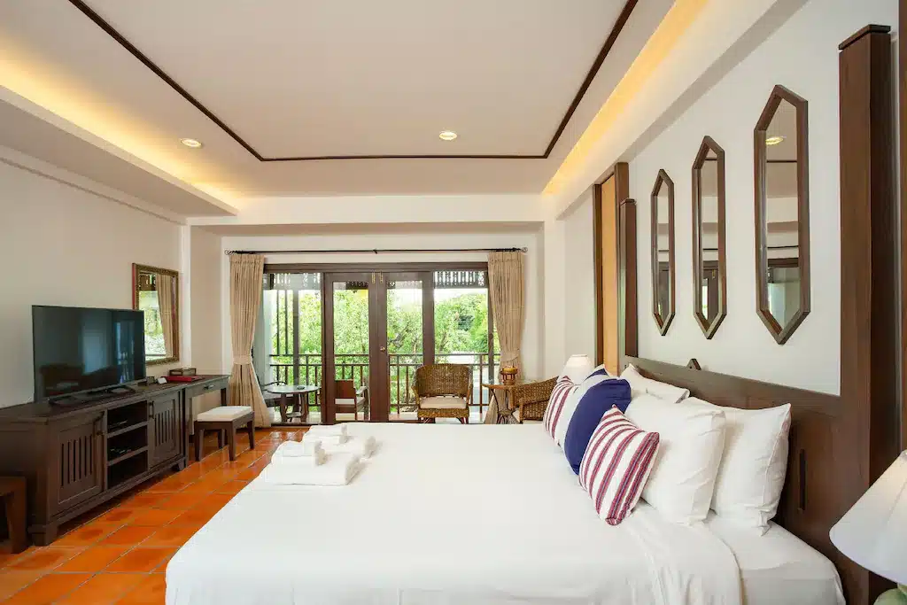 ห้องพักโรงแรมพร้อมเตียงขนาดใหญ่ ทีวี เฟอร์นิเจอร์ไม้ และระเบียงพร้อมเก้าอี้สองตัวที่มองเห็นวิวสีเขียว—เหมาะสำหรับการพักผ่อนหลังจากสำรวจสถานที่ท่องเที่ยวเชียงใหม่.