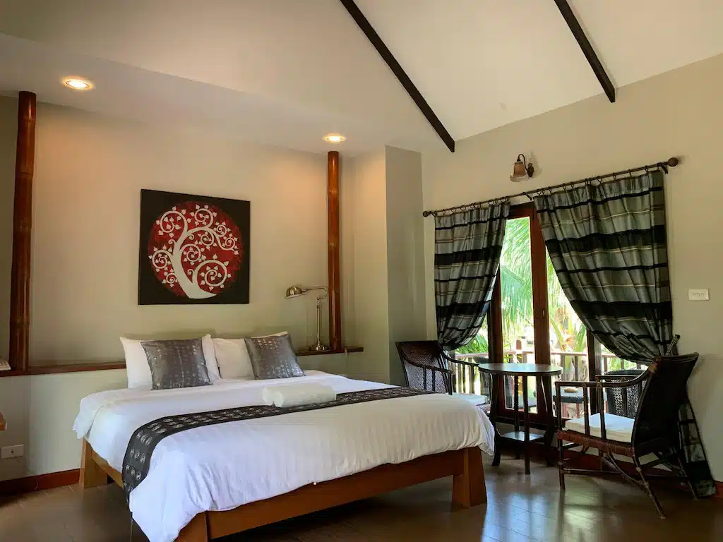 เตียงที่จัดไว้อย่างเรียบร้อยพร้อมหมอนลายและงานศิลปะเหนือเตียง ข้างโต๊ะเล็ก ๆ ที่มีเก้าอี้สองตัวอยู่ใกล้หน้าต่างที่มีผ้าม่านลายทางในห้องนอนที่สว่างและทันสมัยของที่พักริมน้ำอยุธยา.
