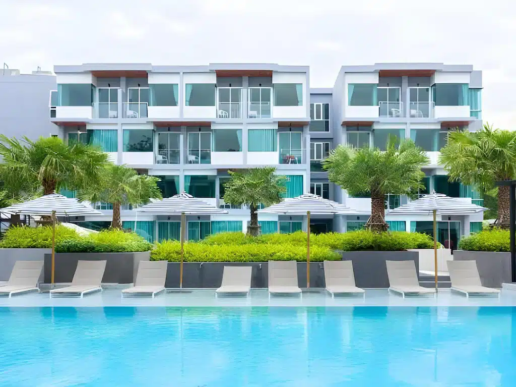 โรงแรมสมัยใหม่พร้อมระเบียงตั้งตระหง่านอยู่ด้านหลังเก้าอี้เลานจ์ ร่ม ต้นปาล์ม และสระว่ายน้ำกลางแจ้งขนาดใหญ่—เหมาะสำหรับผู้ที่มองหาที่พักหัวหินติดทะเลพร้อมบรรยากาศผ่อนคลาย.