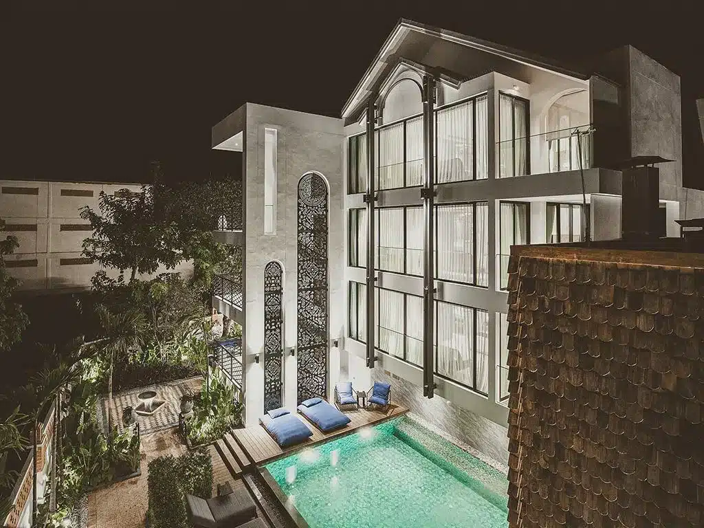 อาคารสามชั้นสมัยใหม่ในยามค่ำคืนพร้อมหน้าต่างบานใหญ่ ภายนอกที่สว่างไสว สระว่ายน้ำ เก้าอี้ชายหาด และสวนที่จัดแต่งอย่างสวยงาม—เหมาะสำหรับผู้ที่มองหาสถานที่ท่องเที่ยวเชียงใหม่ที่ไม่เหมือนใครสำหรับการพักผ่อนครั้งต่อไป.