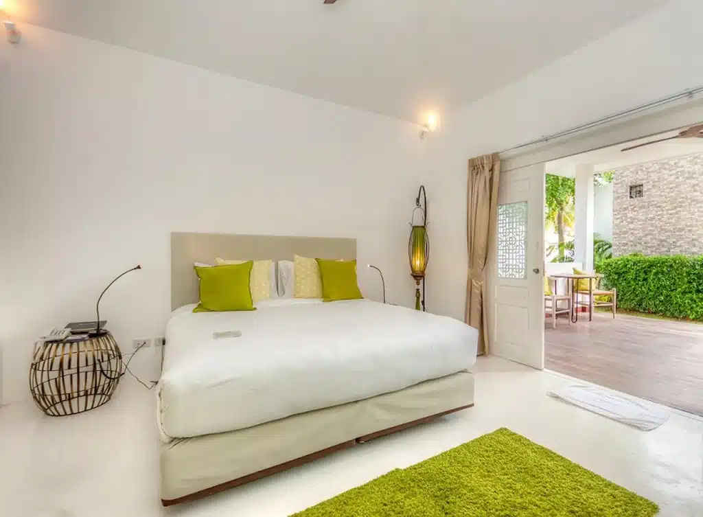 ห้องนอนสไตล์มินิมอลพร้อมเตียงสีขาว หมอนสีเขียว พรมสีเขียว การตกแต่งที่ทันสมัย และประตูเปิดโล่งที่นำไปสู่ลานระเบียงที่มีต้นไม้สีเขียว—เหมาะสำหรับการพักผ่อนที่ โรงแรมหัวหิน หรือ ที่พักหัวหินติดทะเล.