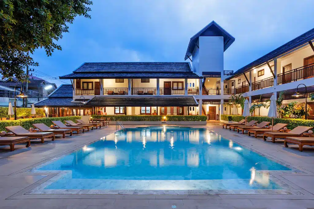 สระว่ายน้ำกลางแจ้งล้อมรอบด้วยเก้าอี้อาบแดด พร้อมอาคารโรงแรมสองชั้นและระเบียงเป็นฉากหลังในยามพลบค่ำ—เหมาะอย่างยิ่งสำหรับการพักผ่อนหลังจากสำรวจสถานที่ท่องเที่ยวในเชียงใหม่.