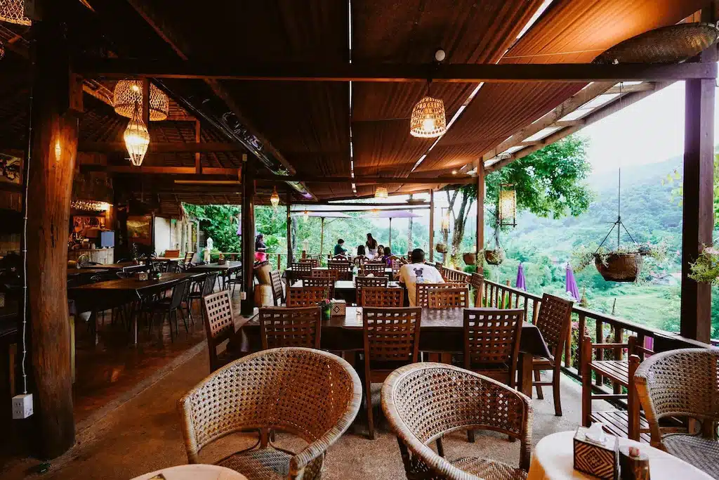 ร้านอาหารกลางแจ้งที่มีเก้าอี้หวายและโต๊ะไม้ มองเห็นวิวธรรมชาติและภูเขาใกล้ที่พักบ่อเกลือ พร้อมด้วยต้นไม้แขวนและผู้คนกำลังรับประทานอาหารเป็นฉากหลัง.