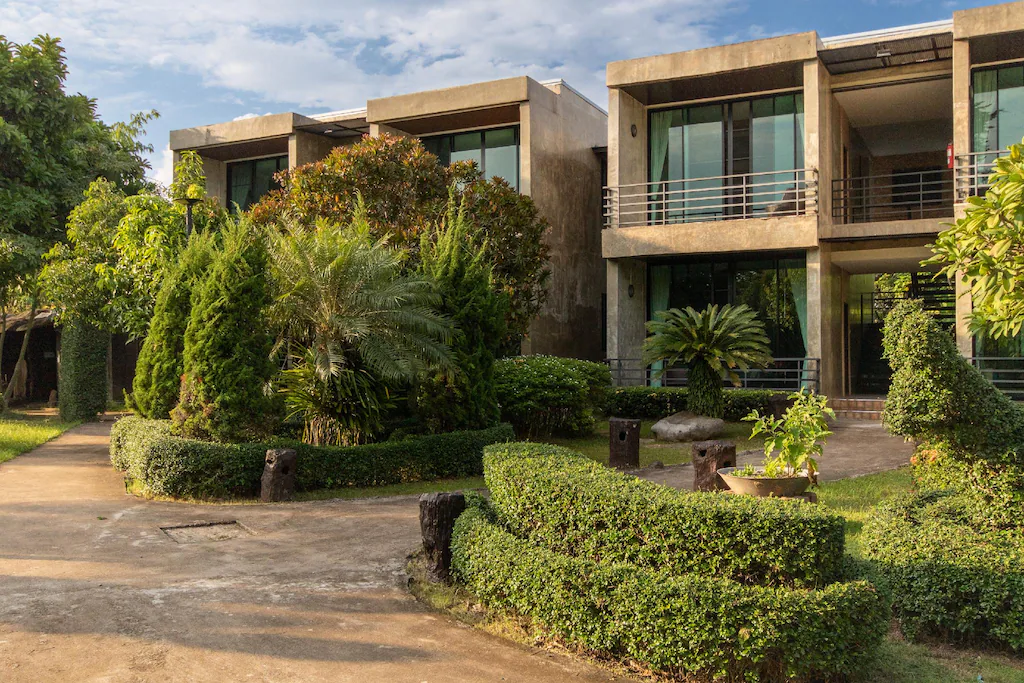 อาคารสองชั้นสมัยใหม่พร้อมหน้าต่างกระจกบานใหญ่ ล้อมรอบด้วยสวนที่ได้รับการดูแลอย่างดีและพุ่มไม้ที่ตัดแต่งอย่างเรียบร้อยในวันที่แดดสดใส—เหมาะสำหรับผู้ที่มองหาที่พักในบรรยากาศเงียบสงบใน น่าน.