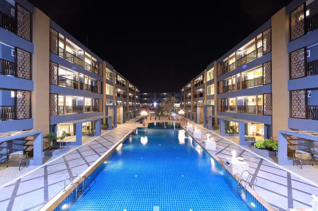 โรงแรมหัวหินที่ทันสมัยและสว่างไสว มีหลายชั้นตั้งอยู่ทั้งสองฝั่งของสระว่ายน้ำรูปสี่เหลี่ยมผืนผ้าขนาดใหญ่ในยามค่ำคืน.