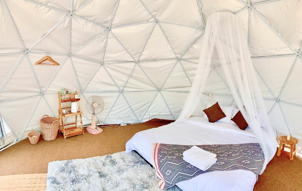 ห้องโดมทรงจีโอเดสิกพร้อมเตียงคู่ใต้มุ้งกันยุง หมอนสีน้ำตาลสองใบ พัดลม ชั้นวางของขนาดเล็ก พรม และตะกร้าบนพื้นพรม—เหมาะอย่างยิ่งสำหรับนักเดินทางที่มองหาที่พักบ่อเกลือที่ไม่เหมือนใครหรือที่พักปัวที่อบอุ่น.