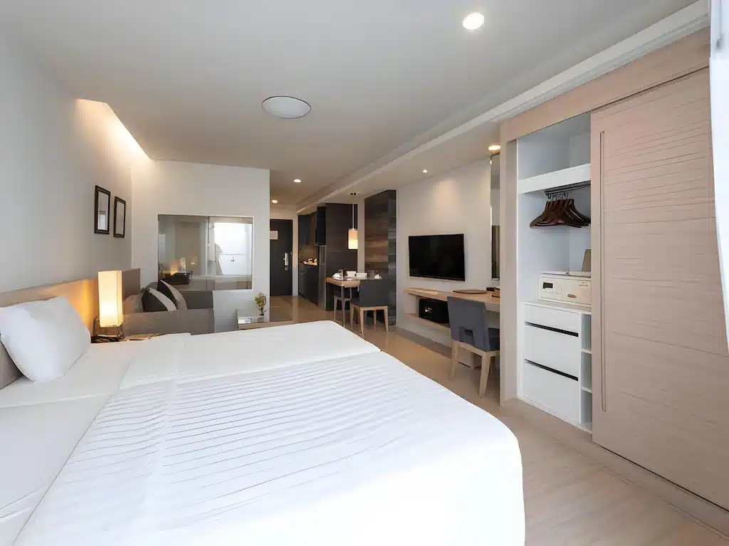 ห้องพักโรงแรมสมัยใหม่พร้อมเตียงขนาดใหญ่ ตู้เสื้อผ้าแบบเปิด ทีวี โต๊ะทำงาน มุมนั่งเล่น และการตกแต่งโทนสีเรียบในแสงไฟนุ่มนวล—เหมาะสำหรับการพักผ่อนหลังจากเที่ยวชมสถานที่ท่องเที่ยวในอยุธยา.