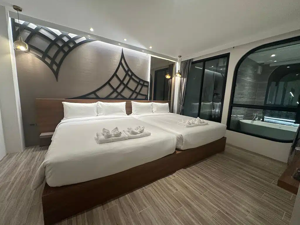 ห้องพักโรงแรมสมัยใหม่พร้อมเตียงคู่สองเตียง ผ้าเช็ดตัวพับไว้ ศิลปะตกแต่งผนัง โคมไฟแขวน พื้นไม้ และหน้าต่างกระจกบานใหญ่ที่มองเห็นห้องน้ำพร้อมอ่างอาบน้ำ—เหมาะสำหรับการเข้าพักของคุณที่ โรงแรมอยุธยา.