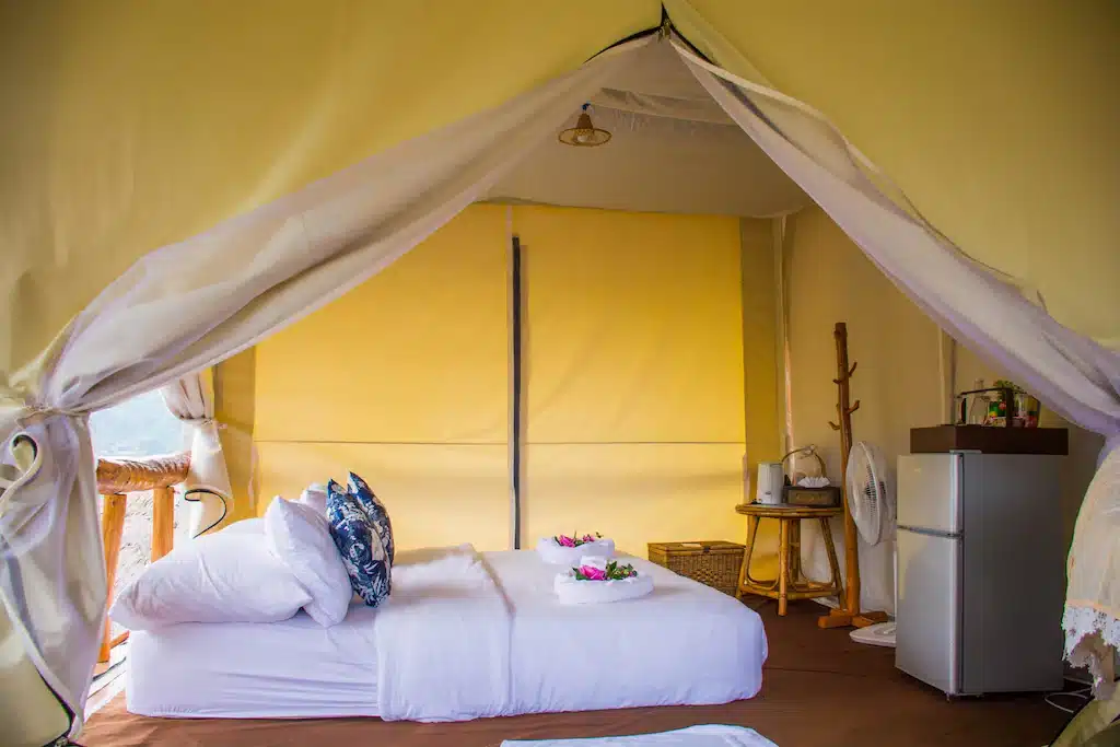 เตียงที่จัดไว้อย่างเรียบร้อยพร้อมผ้าปูที่นอนสีขาวภายในเต็นท์ผ้าใบ ติดกับโต๊ะเล็กๆ ตู้เย็นขนาดเล็ก และสิ่งอำนวยความสะดวกพื้นฐาน—การตั้งแคมป์หรูที่อบอุ่นเหมาะสำหรับผู้ที่มองหาที่พักในบ่อเกลือหรือที่พักในน่านที่เงียบสงบท่ามกลางธรรมชาติ.
