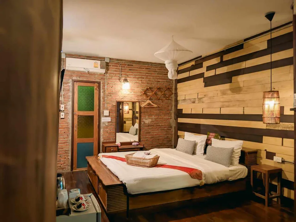 ห้องนอนที่แสนอบอุ่นที่ ที่พักน่าน พร้อมเตียงคู่ ผนังไม้และอิฐ โคมไฟข้างเตียง กระจก และโต๊ะเล็กพร้อมสิ่งอำนวยความสะดวก—เหมาะสำหรับการพักผ่อนหลังจากสำรวจเมือง.