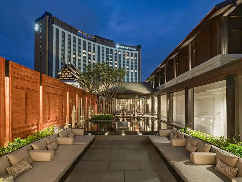 พื้นที่นั่งเล่นกลางแจ้งสมัยใหม่พร้อมเบาะนั่งนุ่ม สระน้ำสะท้อน และอาคารโดยรอบ รวมถึงโรงแรมสูงที่มีโลโก้ InterContinental อยู่ด้านหลังในยามพลบค่ำ—จุดที่น่าดึงดูดสำหรับใครก็ตามที่กำลังมองหาสถานที่ท่องเที่ยวในเชียงใหม่.