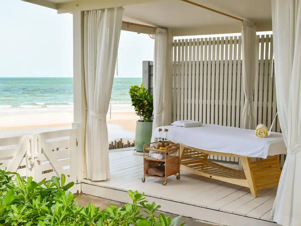 เตียงนวดพร้อมผ้าปูสีขาวและรถเข็นถูกจัดเตรียมไว้ในคาบาน่าที่มองเห็นชายหาด ณ โรงแรมหัวหิน โดยมีคลื่นทะเลเป็นฉากหลังที่สมบูรณ์แบบสำหรับการพักผ่อนริมทะเลของคุณ.