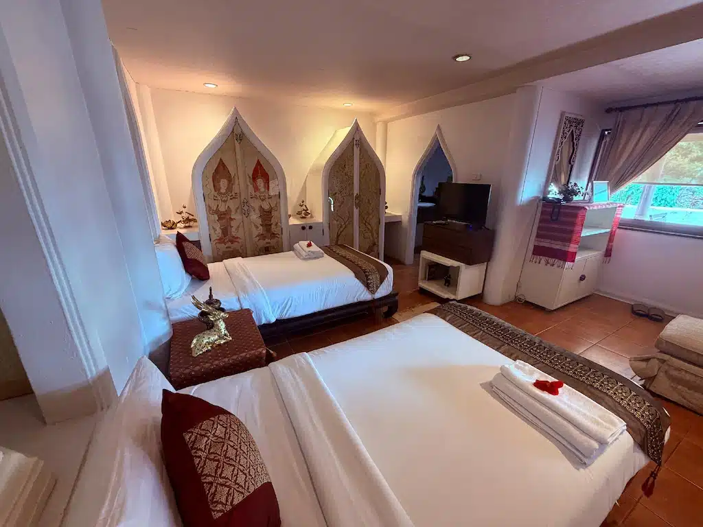 ห้องพักโรงแรมพร้อมเตียงสองเตียง หัวเตียงประดับอย่างหรูหรา ผ้าเช็ดตัวพับเรียบร้อย โทรทัศน์จอแบน และการตกแต่งสไตล์ไทย—เหมาะสำหรับผู้ที่มองหาที่พักอยุธยา หรือโรงแรมอยุธยา ที่มีความสะดวกสบายและสไตล์แท้ๆ.