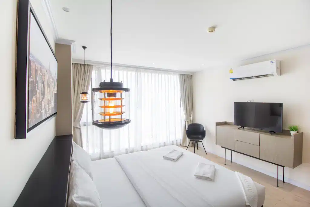 ห้องพักโรงแรมสมัยใหม่ที่ โรงแรมหัวหิน ประกอบด้วยเตียงคู่, โคมไฟข้างเตียง, ศิลปะติดผนัง, โทรทัศน์จอแบนบนตู้, เครื่องปรับอากาศ, และหน้าต่างบานใหญ่พร้อมผ้าม่านสีขาวบางเพื่อการพักผ่อนที่ผ่อนคลายในหัวหิน.