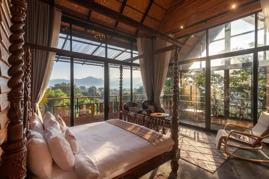 ห้องนอนสไตล์ชนบทที่มีหน้าต่างบานใหญ่ เตียงไม้พร้อมหลังคาเต็นท์ และวิวภูเขา ที่พักในเมืองน่านแห่งนี้เปิดรับแสงธรรมชาติและมองเห็นต้นไม้และพื้นที่นั่งเล่นกลางแจ้งเพื่อการพักผ่อนที่เงียบสงบ.