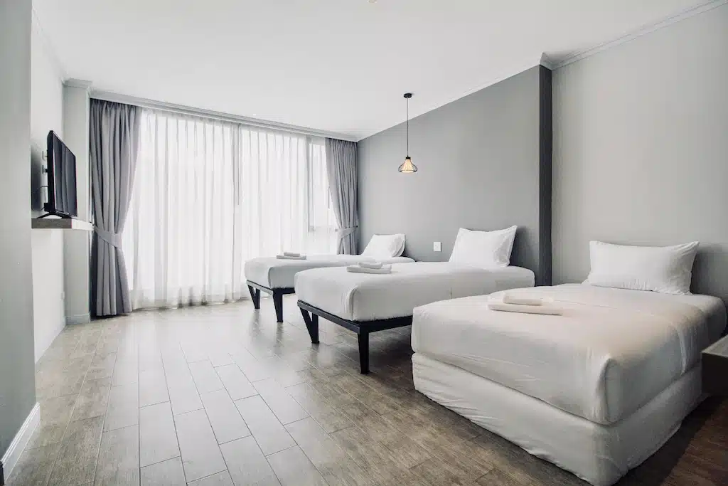 ห้องพักโรงแรมสมัยใหม่ที่ ที่พักหัวหิน พร้อมเตียงเดี่ยวสามเตียง ผ้าปูที่นอนสีขาว โทรทัศน์ติดผนัง หน้าต่างบานใหญ่พร้อมผ้าม่านบาง และพื้นไม้—เหมาะสำหรับการพักผ่อนริมทะเลของคุณ.