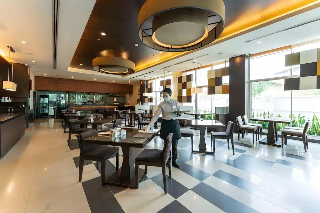 พนักงานเสิร์ฟสวมหน้ากากถือถาดในร้านอาหารสมัยใหม่ที่มีแสงสว่างเพียงพอ พร้อมโต๊ะที่จัดเรียงอย่างเรียบร้อยและการตกแต่งที่มีรูปทรงเรขาคณิต มอบประสบการณ์การรับประทานอาหารที่หรูหราให้กับแขกที่ที่พักอยุธยา.