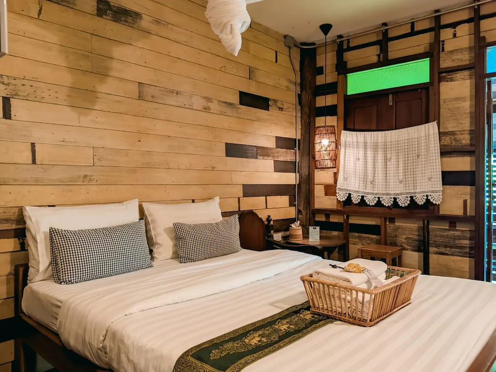 ห้องนอนที่อบอุ่นพร้อมเตียงคู่ ผนังไม้แผ่น กำแพงตกแต่งด้วยหมอน และตะกร้าผ้าเช็ดตัวบนเตียง รอคุณอยู่ที่ ที่พักน่ารักในเมืองน่าน พร้อมหน้าต่างที่ตกแต่งด้วยผ้าม่านลูกไม้.