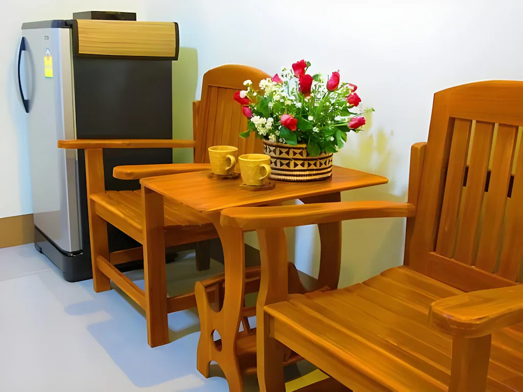 เก้าอี้ไม้สองตัวและโต๊ะเล็ก ๆ พร้อมแก้วและดอกไม้ตั้งอยู่ข้างตู้เย็นในห้องที่สว่างไสว—เหมาะสำหรับการผ่อนคลายที่ ที่พักน่าน หรือ ที่พักปัว ที่แสนสบายของคุณ.