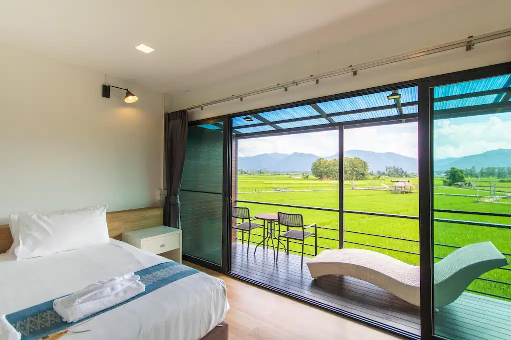 ห้องนอนสมัยใหม่ที่ ที่พักปัว พร้อมเตียงขนาดใหญ่ ประตูกระจกเปิดออกสู่ระเบียงที่มีโต๊ะ เก้าอี้ เก้าอี้เลานจ์ และวิวทิวทัศน์ที่สวยงามของทุ่งหญ้าเขียวขจีและภูเขาไกล.