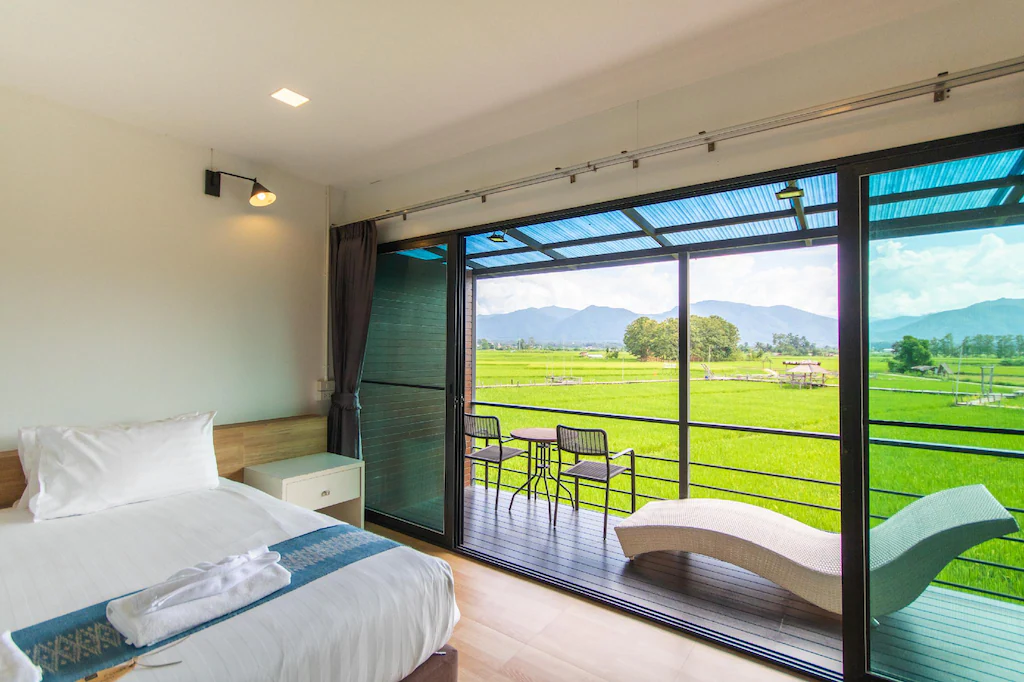 ห้องพักโรงแรมสมัยใหม่พร้อมเตียง, โต๊ะข้างเตียง, และเก้าอี้พักผ่อน; ประตูบานเลื่อนกระจกเปิดออกสู่ระเบียงที่มีเก้าอี้สำหรับชมวิวทุ่งหญ้าเขียวขจีและภูเขา—เหมาะสำหรับการพักผ่อน ที่พักปัว หรือ ที่พักน่าน.