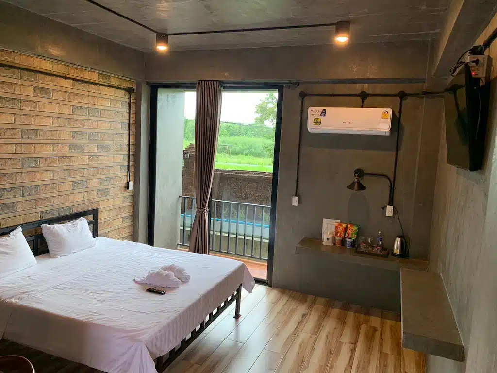 ห้องนอนสมัยใหม่ที่โรงแรมอยุธยา พร้อมเตียงคู่ ผนังอิฐและคอนกรีต ทีวีติดผนัง เครื่องปรับอากาศ ของว่างบนชั้นวาง และหน้าต่างที่มองเห็นวิวสีเขียว.