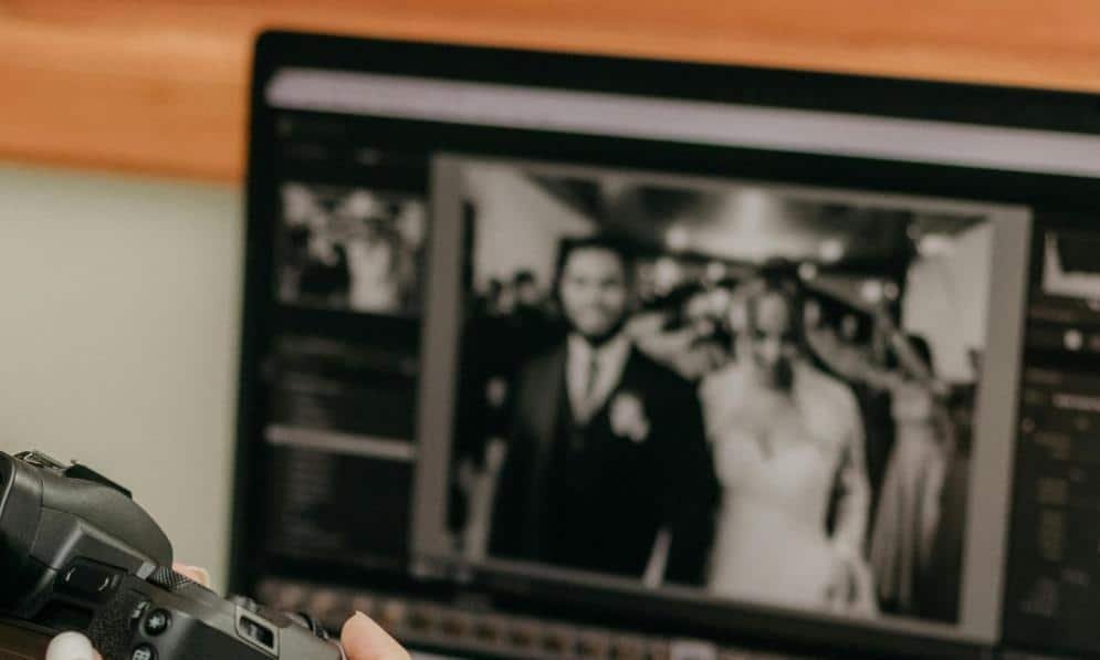 บุคคลหนึ่งกำลังแก้ไขรูปถ่ายงานแต่งงานที่เบลอของเจ้าสาวและเจ้าบ่าวบนหน้าจอคอมพิวเตอร์ ขณะถือกล้องอยู่.