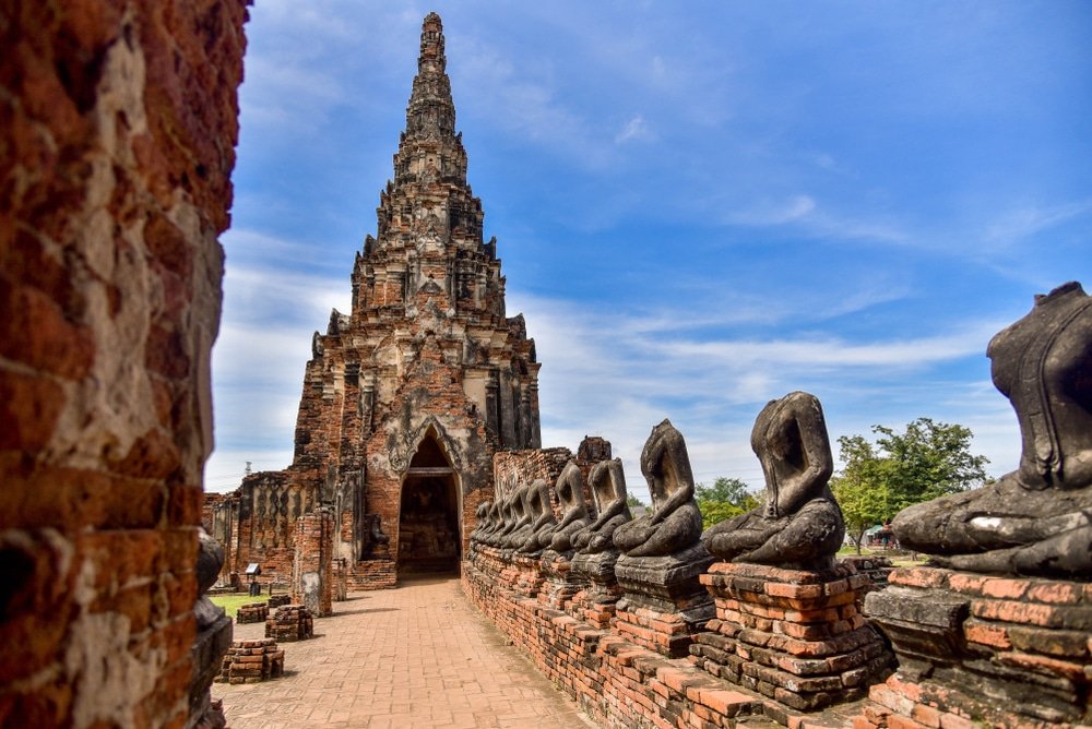 วัดโบราณก่ออิฐพร้อมยอดแหลมสูง ล้อมรอบด้วยพระพุทธรูปไร้ศีรษะบนกำแพงอิฐ ท่ามกลางท้องฟ้าสีคราม—หนึ่งในสถานที่ท่องเที่ยวอยุธยาที่ต้องไปเยือนสำหรับผู้รักประวัติศาสตร์.